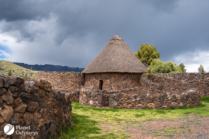 The historical site of Raqchi in Peru, features ancient Inca stone walls and a reconstructed thatched-roof hut, surrounded by green landscapes and Andean mountains